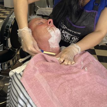 A straight-razor shave in progress, lather along the jaw and a warm towel across the chest.