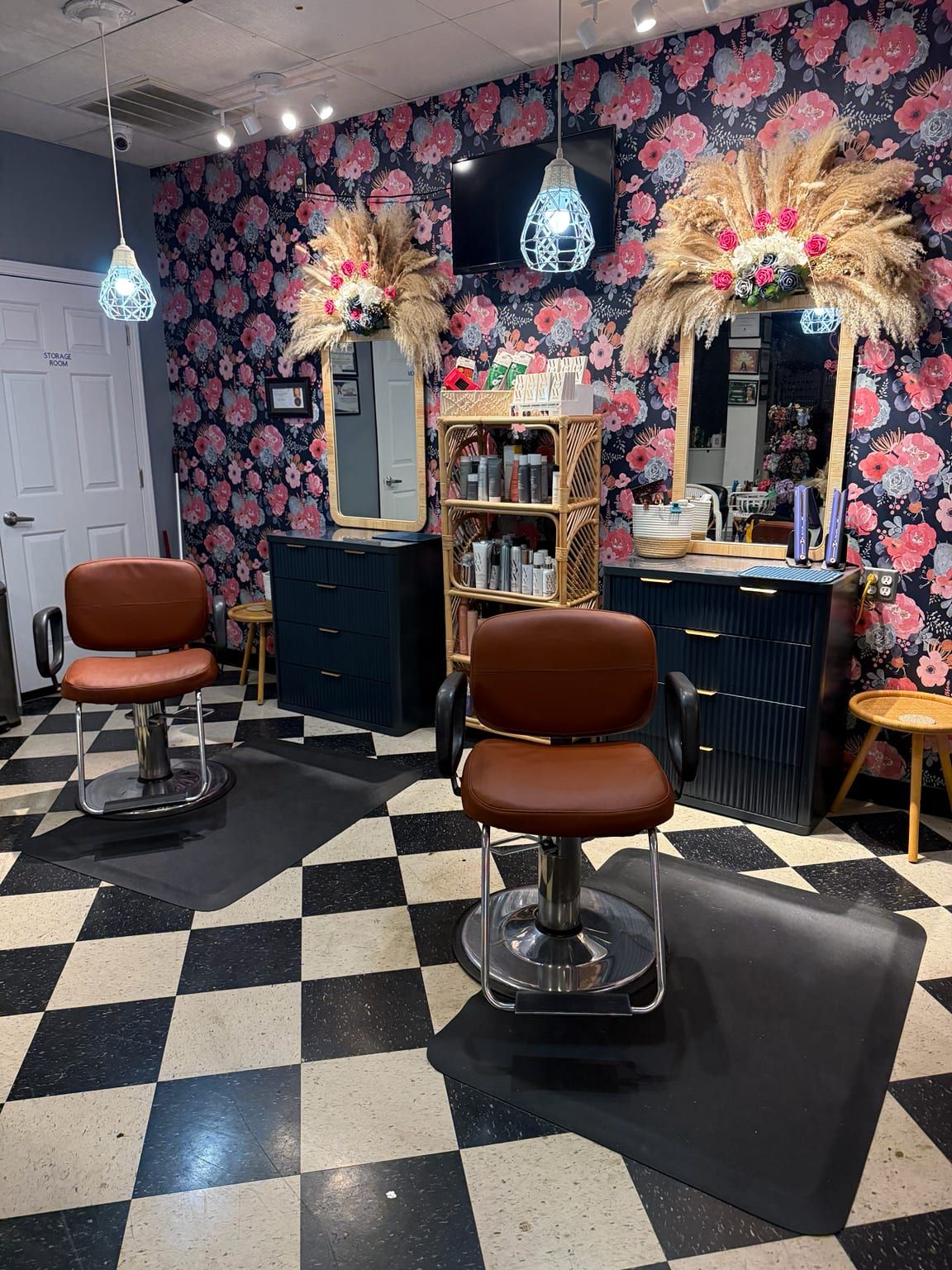 Two styling chairs on a black and white checkerboard floor, facing mirrors set into dark floral wallpaper, with pampas grass above them.
