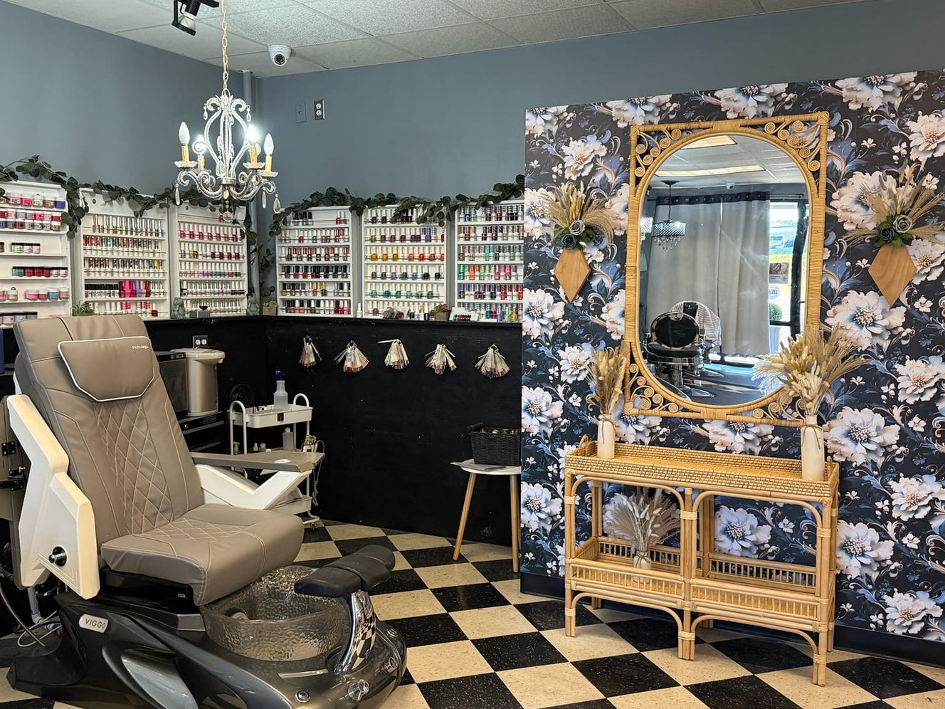A pedicure chair beside a wall of nail polish on white shelves, and an arched wicker mirror against dark floral wallpaper.